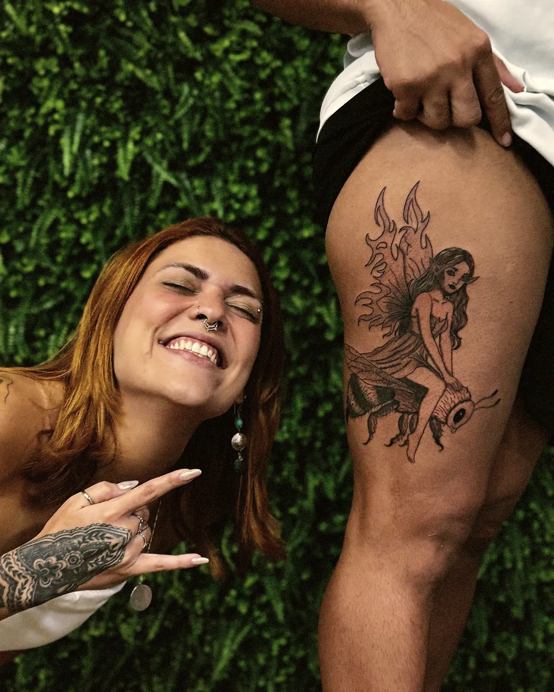 Aluna sorrindo ao lado da tatuagem finalizada na coxa, fundo de folhagem da escola