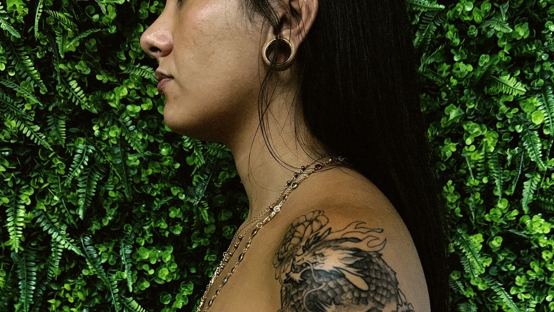 Aluna de perfil mostrando tatuagem em preto e cinza no braço, fundo de folhagem