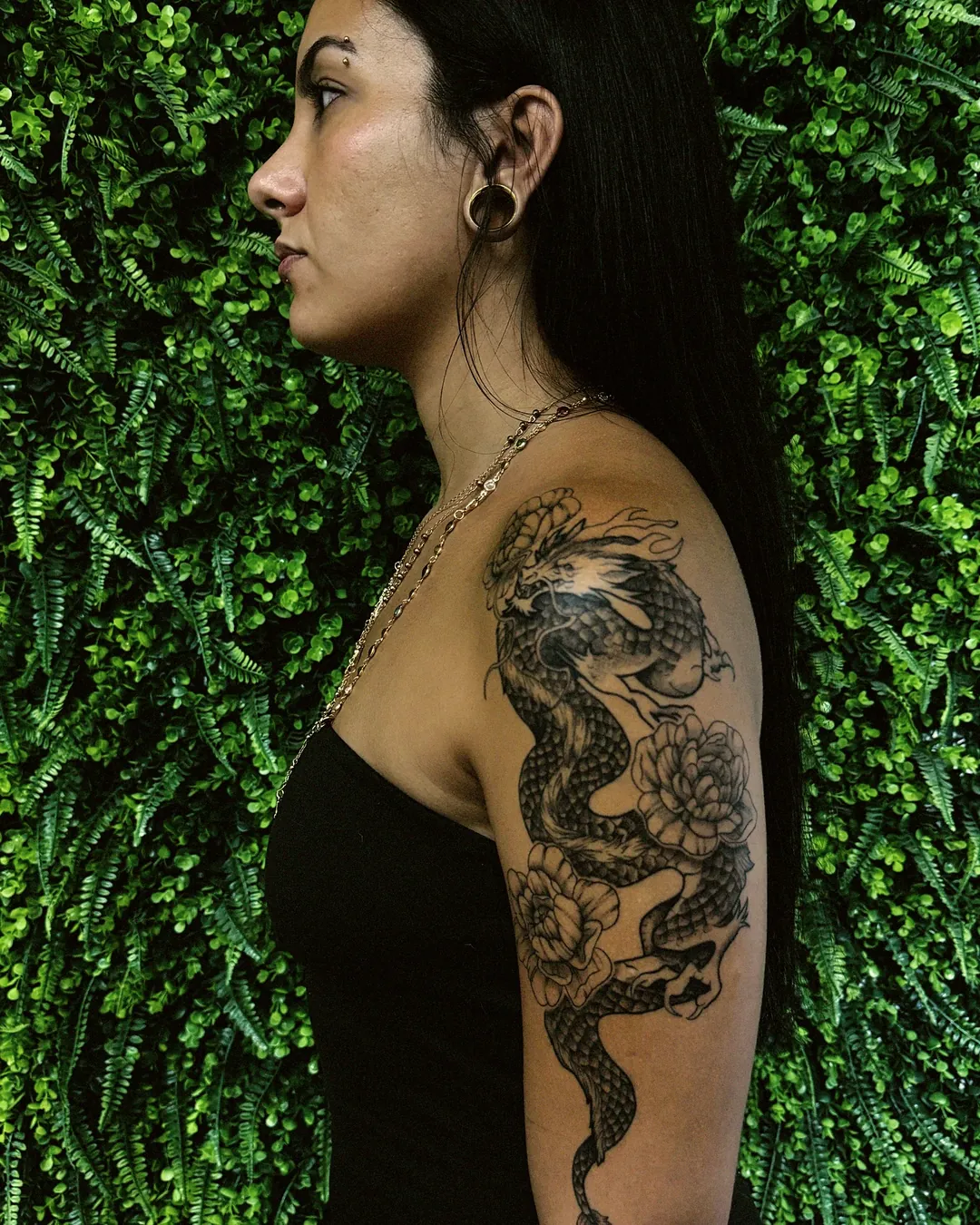 Aluna de perfil mostrando tatuagem em preto e cinza no braço, fundo de folhagem