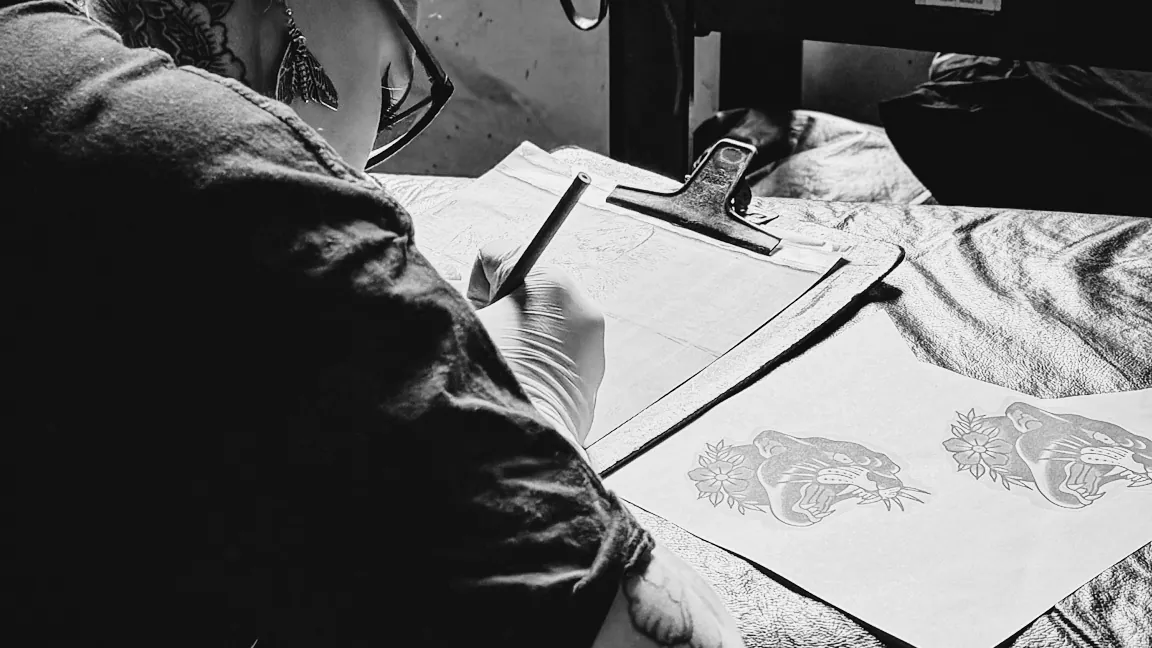 Tatuadora ruiva desenhando o projeto da tatuagem na mesa ao lado da janela da escola The VOID