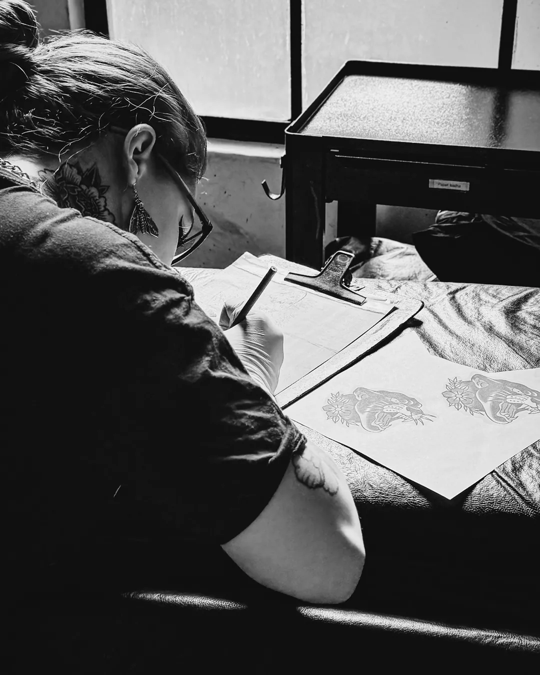 Tatuadora ruiva desenhando o projeto da tatuagem na mesa ao lado da janela da escola The VOID